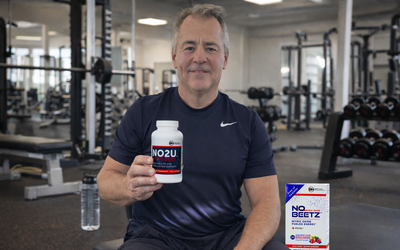 Dr. Nathan S Bryan in a gym holding NO2U nitric oxide lozenges, highlighting nitric oxide support for circulation, cardiovascular health, energy, and active aging in a fitness lifestyle setting with N.O. Beetz sitting on the bench.