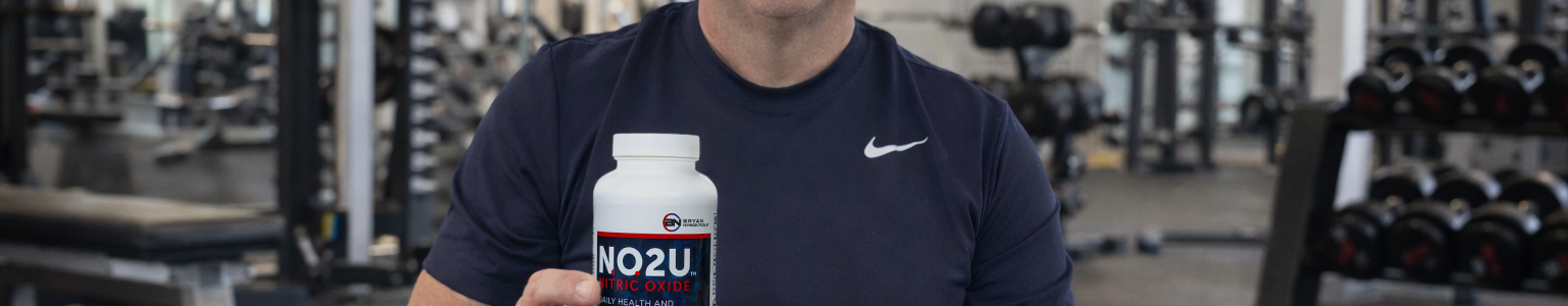 Dr. Nathan S Bryan in a gym holding NO2U nitric oxide lozenges, highlighting nitric oxide support for circulation, cardiovascular health, energy, and active aging in a fitness lifestyle setting with N.O. Beetz sitting on the bench.