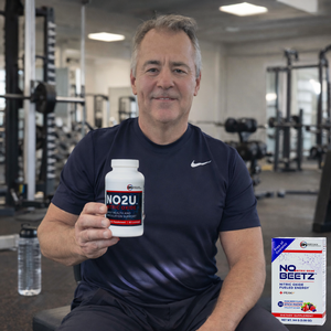 Dr. Nathan S Bryan in a gym holding NO2U nitric oxide lozenges, highlighting nitric oxide support for circulation, cardiovascular health, energy, and active aging in a fitness lifestyle setting with N.O. Beetz sitting on the bench.