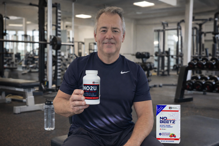 Dr. Nathan S Bryan in a gym holding NO2U nitric oxide lozenges, highlighting nitric oxide support for circulation, cardiovascular health, energy, and active aging in a fitness lifestyle setting with N.O. Beetz sitting on the bench.