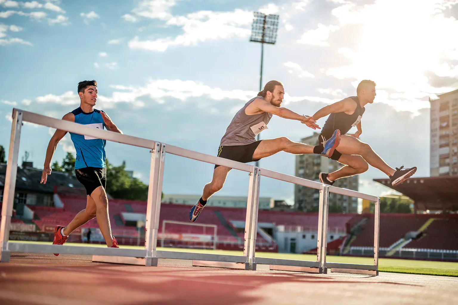 Track athletes sprinting and clearing hurdles on an outdoor stadium track, showcasing speed, endurance, and peak performance supported by nitric oxide for improved blood flow, oxygen delivery, and athletic power.