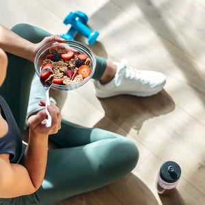Active woman eating a healthy post-workout bowl with berries and whole grains on the gym floor, highlighting nutrition that supports nitric oxide production, blood circulation, recovery, and overall fitness performance.