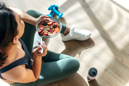 Active woman eating a healthy post-workout bowl with berries and whole grains on the gym floor, highlighting nutrition that supports nitric oxide production, blood circulation, recovery, and overall fitness performance.