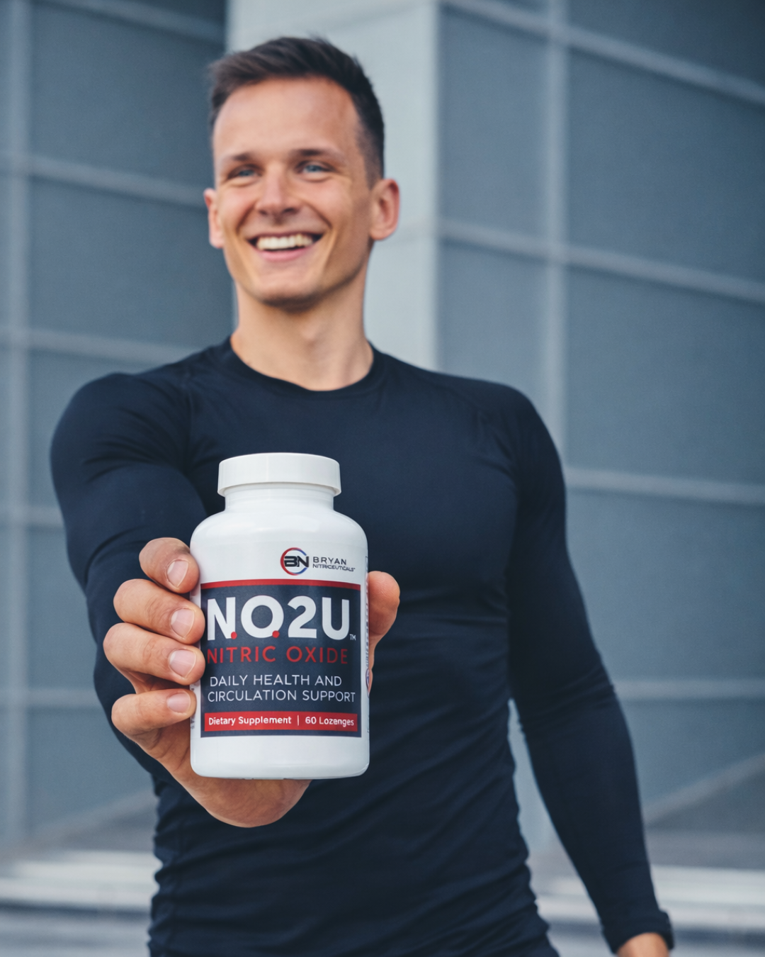 Man holding a supplement bottle labeled 'NO2U' in front of a modern building.